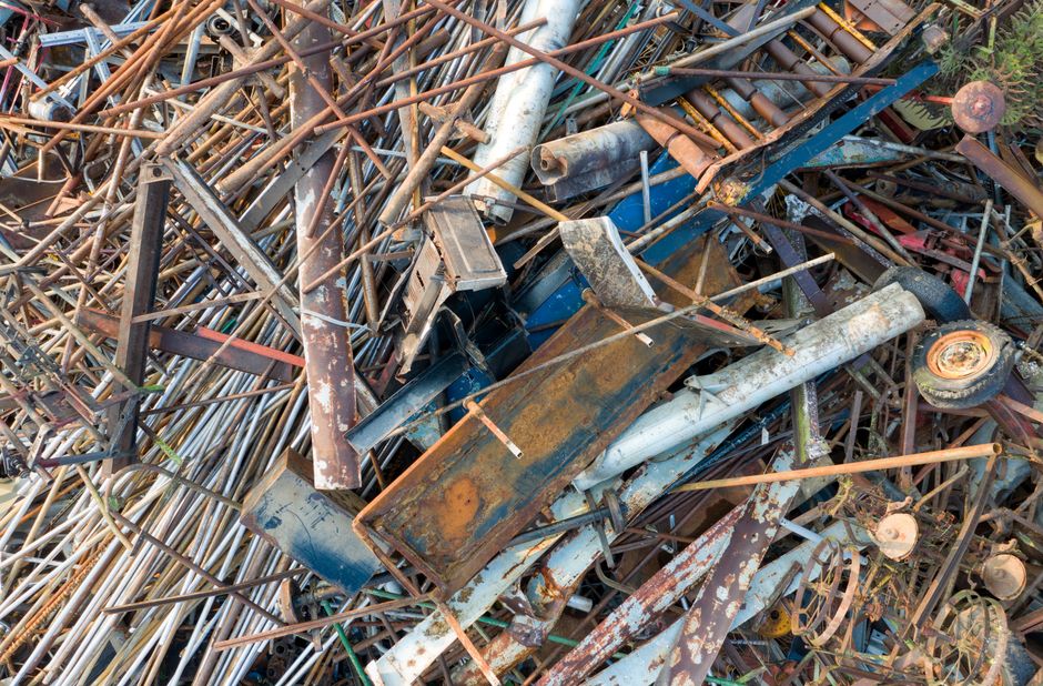 A large pile of assorted scrap metal, including rusted pipes, broken metal sheets, and discarded machinery parts, is situated outdoors on a pile of thin metal rods and framework. Some objects are rust