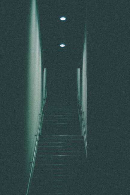 Concrete outdoor staircase in a narrow alleyway leading up to residential buildings, flanked by a black metal handrail on the left and a weathered metal pole on the right. The steps are slightly worn