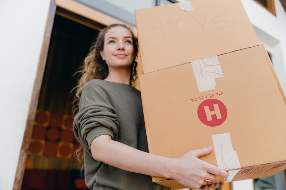 A young woman with long, curly hair and a calm expression is standing indoors, holding a stack of three cardboard boxes during a home relocation. The top box is labeled 'toys' in handwritten letters a