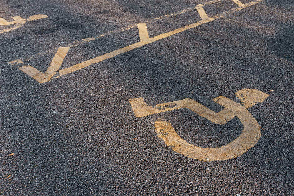 Close-up photograph of a designated parking space for disabled persons on an asphalt surface, with faded yellow lines and a wheelchair symbol painted on the ground. The parking space is situated near