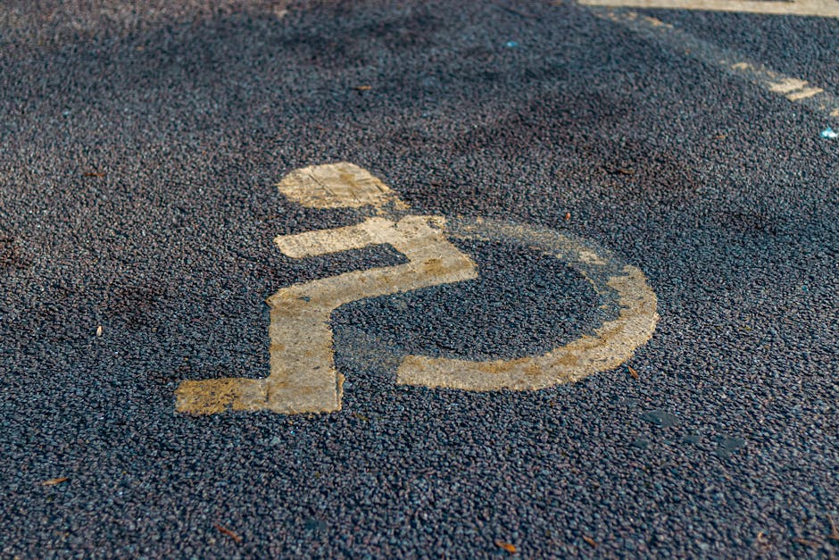 Close-up photograph of a designated parking space for disabled persons on an asphalt surface, with faded yellow lines and a wheelchair symbol painted on the ground. The parking space is situated near